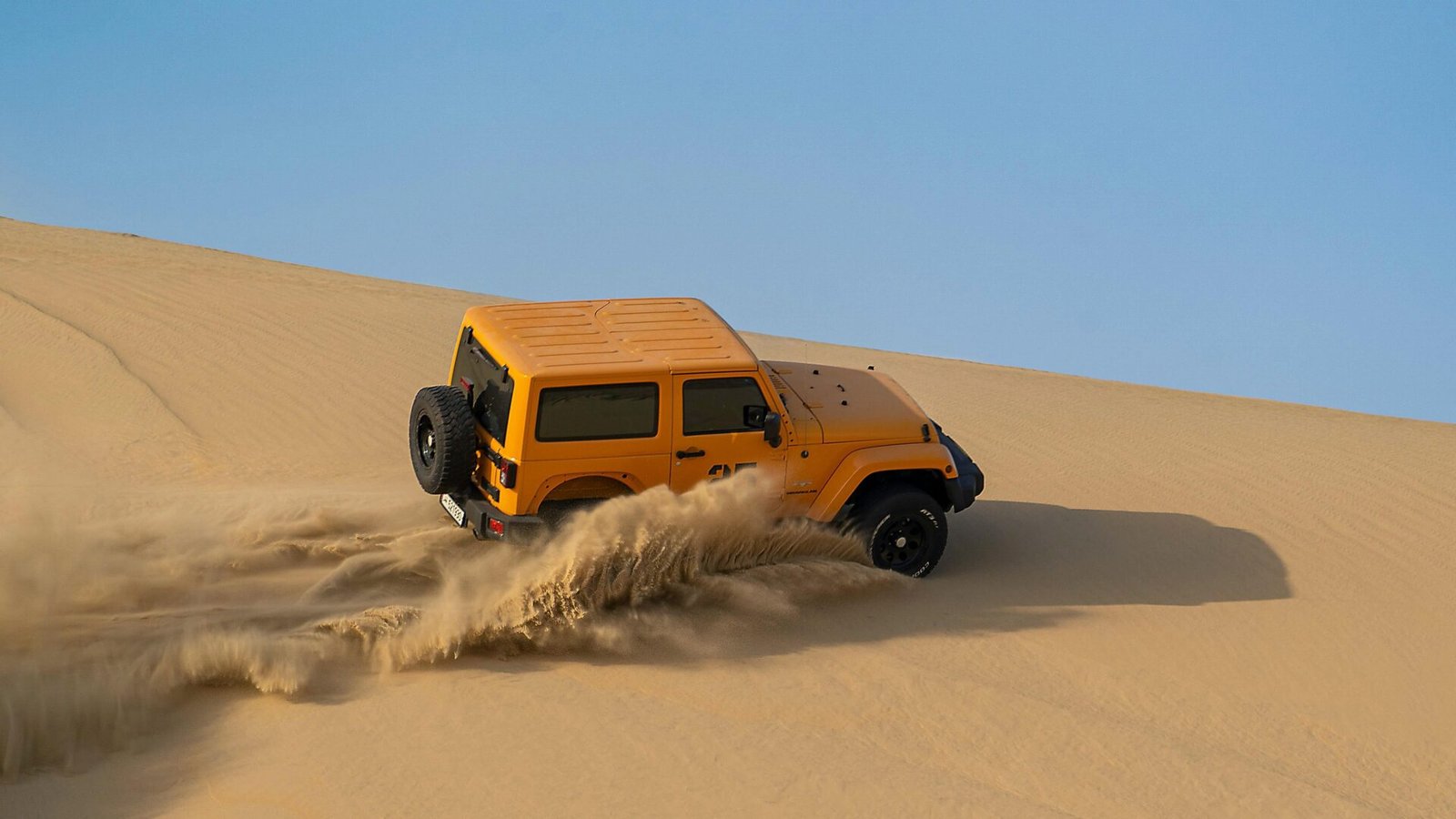 Yellow 4x4 with a spare wheel on the rear door climbing a steep Namib dune, throwing up a wall of sand