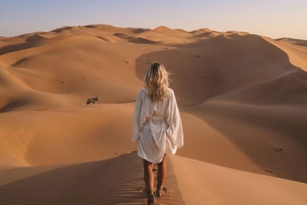 Woman in a white shirt walking a dune ridge in the Namib at sunrise, a lone oryx on the sand behind her