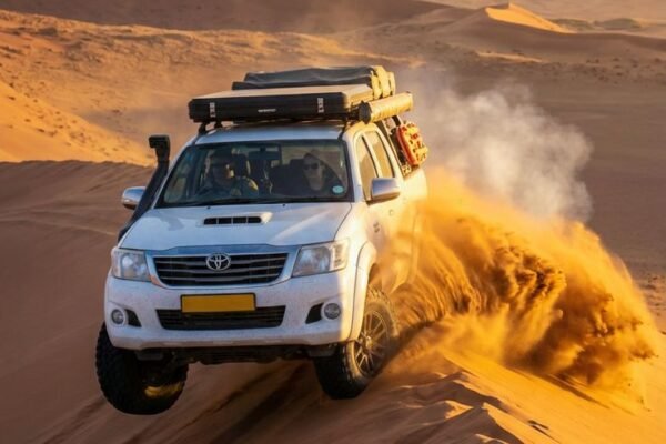 White Toyota Hilux with a roof rack driving along a dune crest in the Namib, spraying golden sand