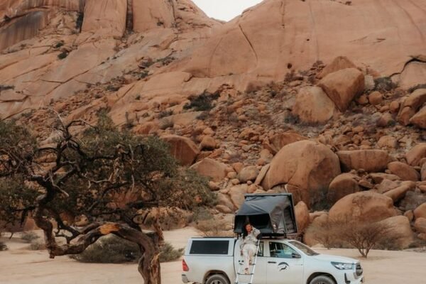 4x4 with a rooftop tent open, parked under a tree beside a stone picnic bench among the Spitzkoppe boulders