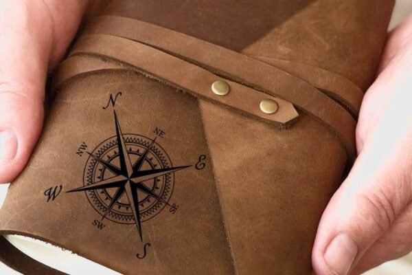 Hands holding a leather-bound travel journal with a compass rose embossed on the cover and a wrap-around tie