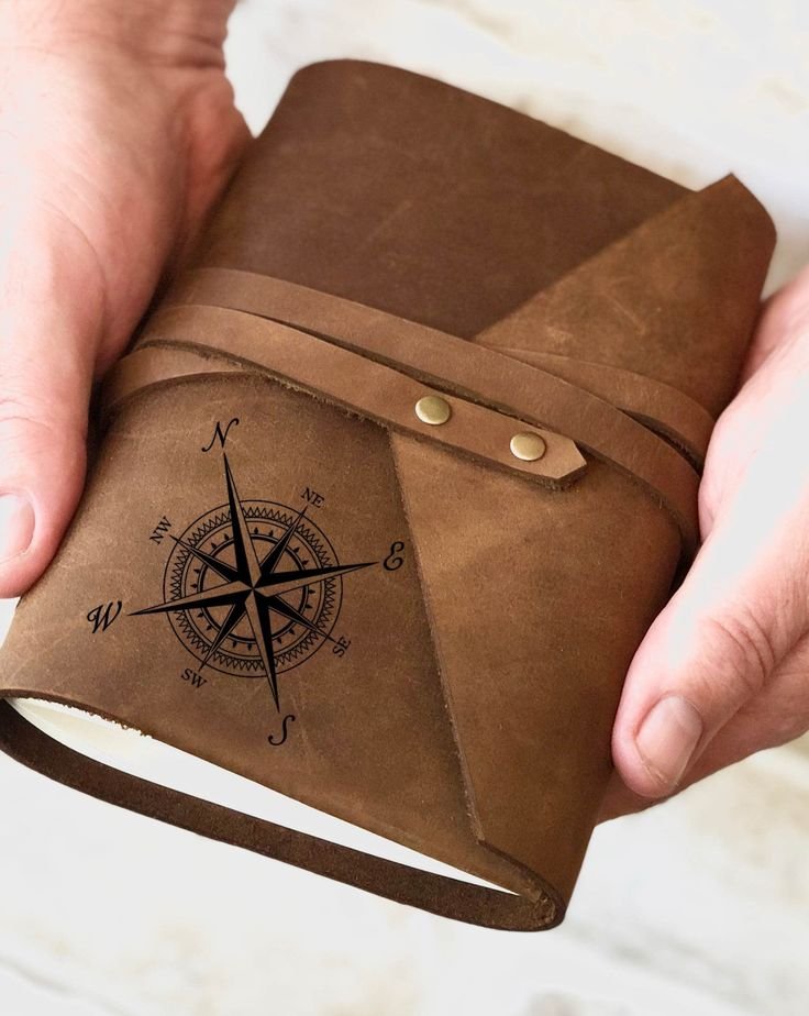 Hands holding a leather-bound travel journal with a compass rose embossed on the cover and a wrap-around tie