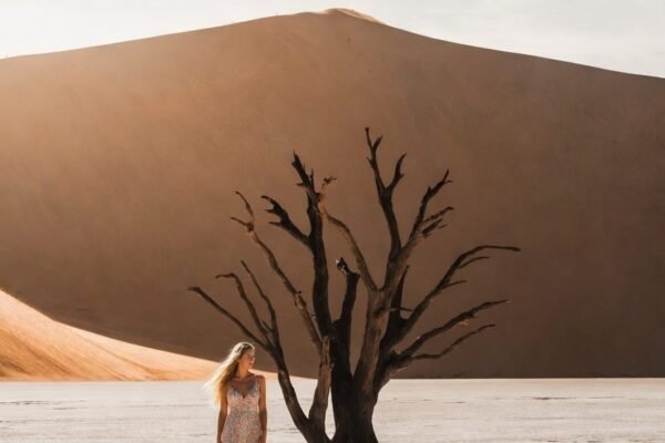 Woman in a pale dress walking towards a dead camelthorn tree on the white clay pan at Deadvlei, a red dune rising behind