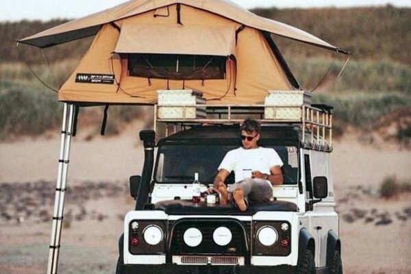Traveller sitting on the bonnet of a Land Rover Defender with a rooftop tent open, parked on the coast