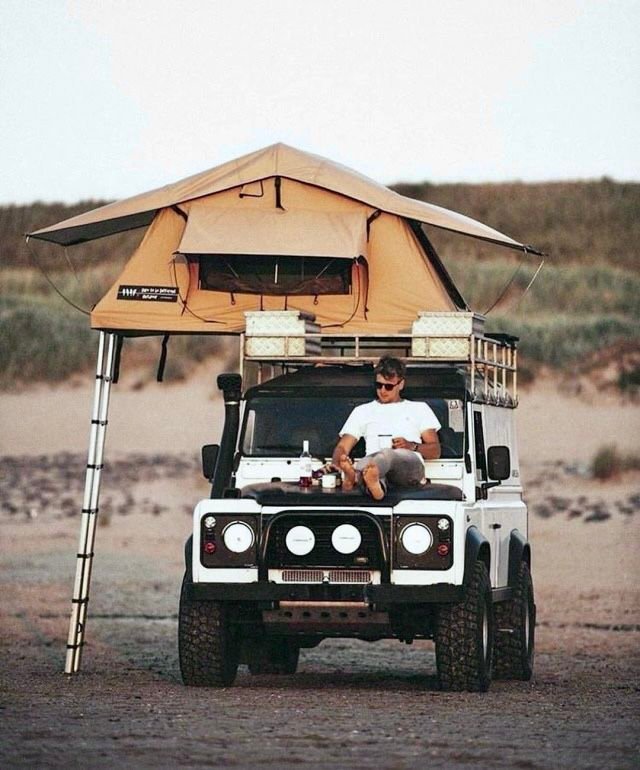 Traveller sitting on the bonnet of a Land Rover Defender with a rooftop tent open, parked on the coast