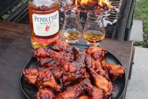 Barbecued chicken pieces on a plate beside a braai fire, with a bottle of whisky and two glasses on the table