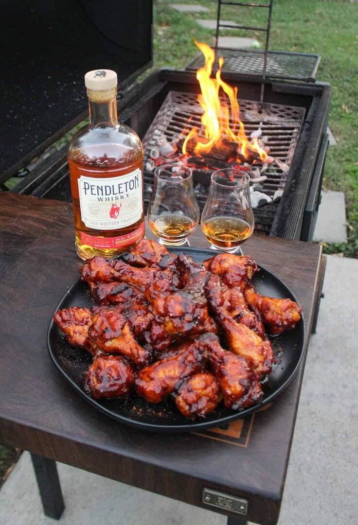 Barbecued chicken pieces on a plate beside a braai fire, with a bottle of whisky and two glasses on the table