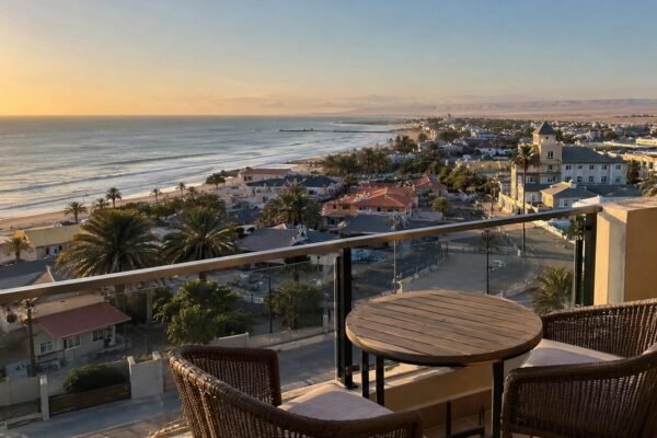 Swakopmund beachfront at sunset seen from a balcony, with palm-lined streets, the jetty and the Namib dunes meeting the Atlantic behind the town